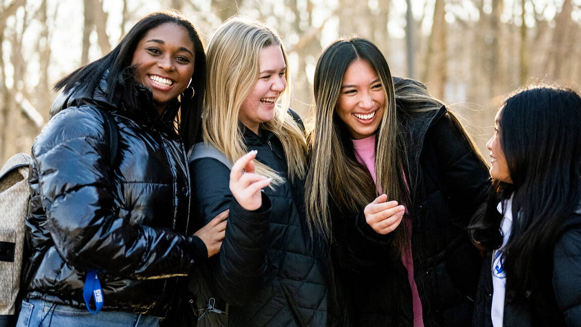 Four individuals laugh together outdoors in winter attire, surrounded by bare trees and snow.